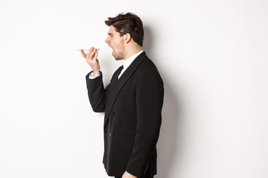 Profile shot of angry businessman in black suit, shouting at speakerphone and looking mad, recording voice message, standing over white background.