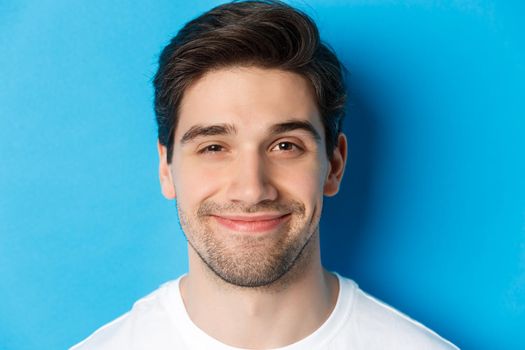 Headshot of attractive man smiling pleased, looking intrigued, standing over blue background.