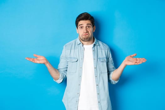 Confused young man in casual clothes shrugging, dont know anything, looking clueless, standing against blue background.