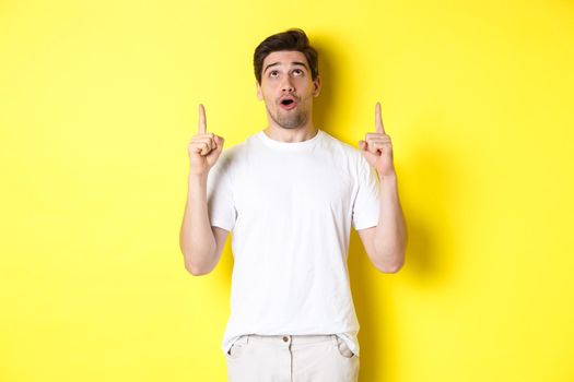 Impressed guy checking out promo offer, looking and pointing fingers up at advertisement, standing over yellow background.