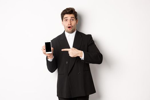 Portrait of attractive businessman in black suit, looking surprised and pointing finger at smartphone sreen, standing over white background.