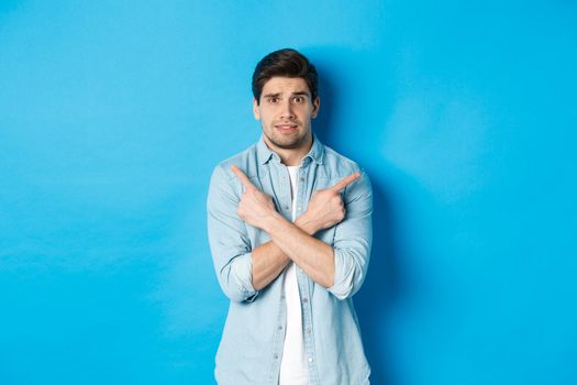 Portrait of nervous guy pointing fingers sideways, looking indecisive and asking for help with choice, showing left and right promo offers, standing against blue background.