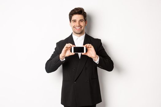 Portrait of attractive businessman in black suit, holding smartphone horizontally and showing screen, smiling pleased, standing against white background.