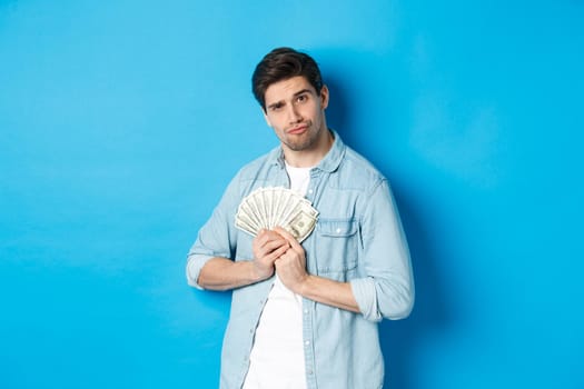 Handsome young man keeping money to himself, smiling and looking greedy, standing over blue background.