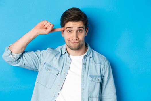 Close-up of man scolding for acting stupid or crazy, rolling finger on head and looking at camera, standing over blue background.