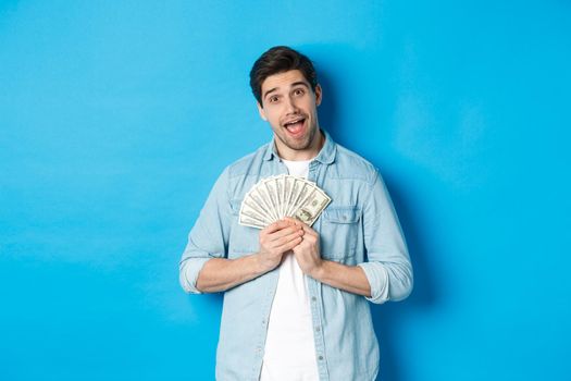 Smiling greedy guy hugging money and smiling, unwilling to share, standing over blue background.