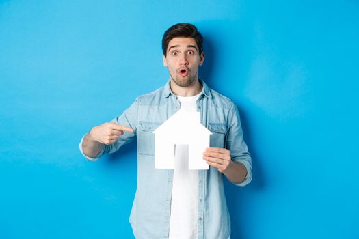 Insurance, mortgage and real estate concept. Surprised young man pointing at house card model and looking at camera, standing against blue background.