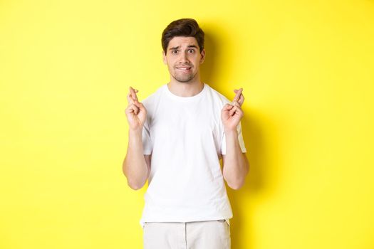 Nervous guy crossing fingers for good luck, hoping for something, standing over yellow background.