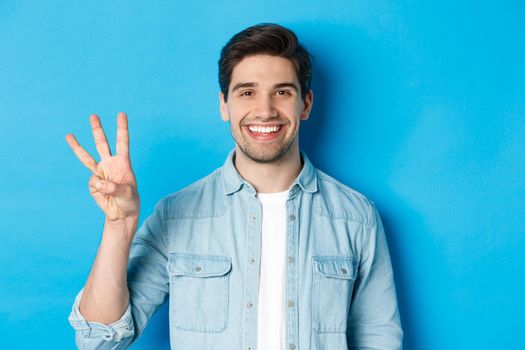 Close-up of handsome man smiling, showing fingers number three, standing over blue background.