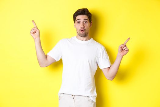 Handsome man pointing fingers sideways, showing two promos, standing over yellow background.