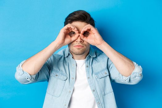 Close-up of man holding hands on eyes, looking through binoculars with serious face, standing over blue background.