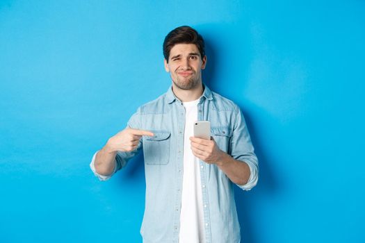 Concept of online shopping, applications and technology. Skeptical and displeased guy pointing finger at smartphone and grimacing disappointed, standing over blue background.