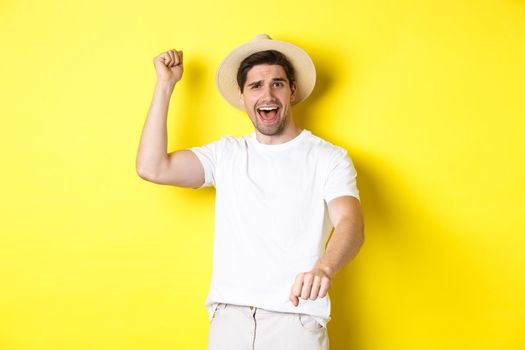 Concept of tourism and summer. Young man traveller showing rodeo gesture, standing in straw hat and white clothes, standing over yellow background.