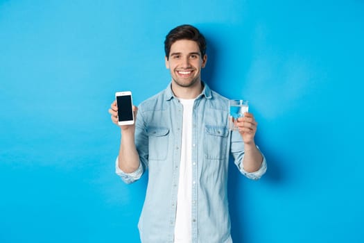 Young man control water balance with smartphone app, showing mobile screen app and smiling, standing over blue background.