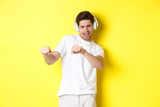 Cool guy listening music in headphones and dancing, standing in white clothes against yellow studio background.