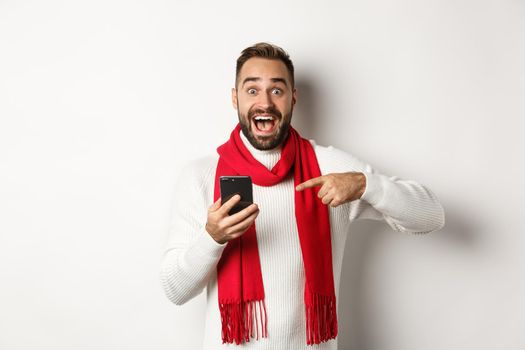 Excited man telling about offer on mobile phone, pointing at smartphone and looking amazed, standing against white background.