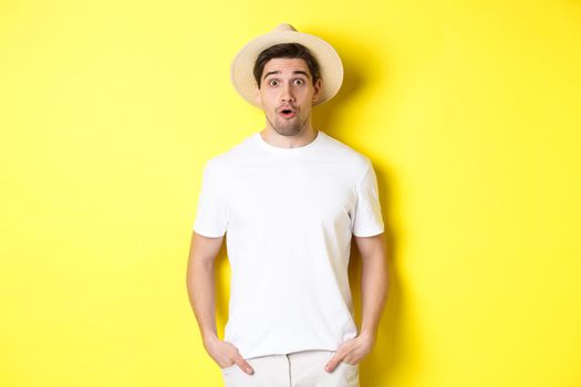 Concept of travelling and tourism. Surprised caucasian guy in straw hat, say wow, standing against yellow background.