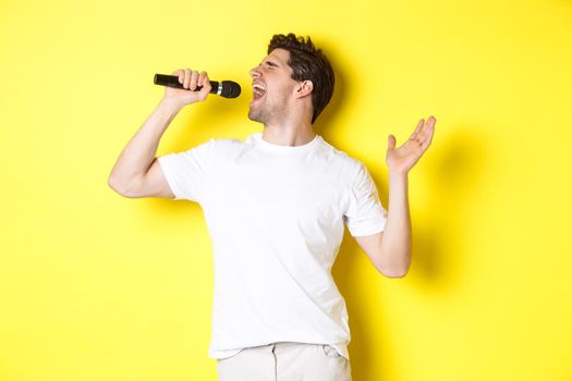 Young man singer holding microphone, reaching high note and singing karaoke, standing over yellow background.