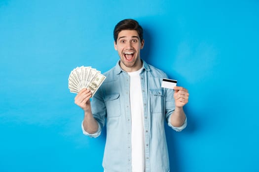 Happy attractive man looking amazed, showing cash and credit card, concept of banks, credit and finance. Blue studio background.