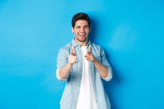 Cheeky handsome guy pointing fingers at you, winking flirty, standing in casual outfit against blue background.