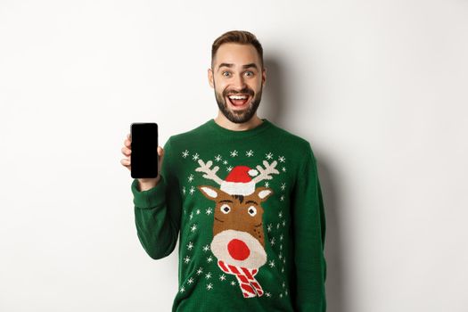 New Year, holidays and celebration. Excited bearded man in Christmas sweater, showing smartphone screen, standing over white background.
