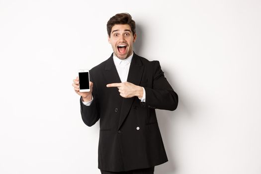 Portrait of handsome businessman in suit, pointing finger at mobile phone screen, showing advertisement, standing over white background.