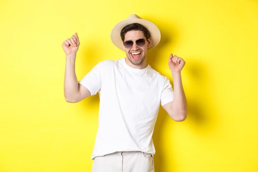 Tourism, travelling and holidays concept. Happy caucasian guy dancing and having fun on vacation, wearing sunglasses with straw hat, standing against yellow background.