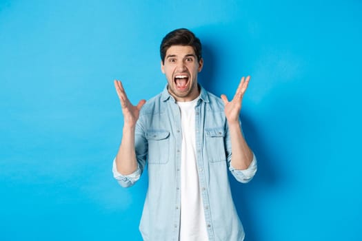 Frustrated young man screaming and looking tensed, shaking hands mad, standing against blue background.