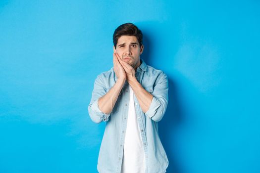 Portrait of man with toothache grimacing from pain and touching cheek, standing against blue background.