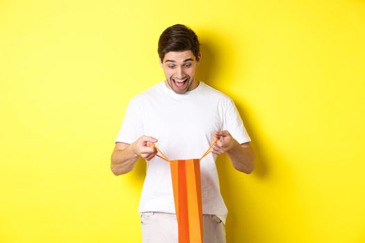 Excited guy open bag with gift, looking inside with amazement and happy face, standing against yellow background.