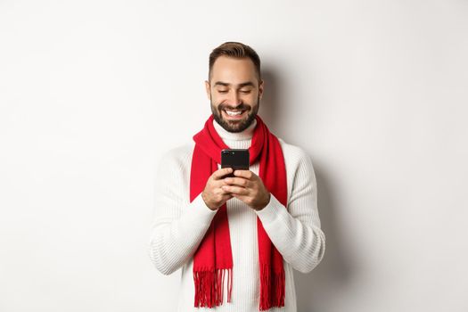 Happy man smiling while reading message on mobile phone, standing in winter sweater and red scarf against white background.