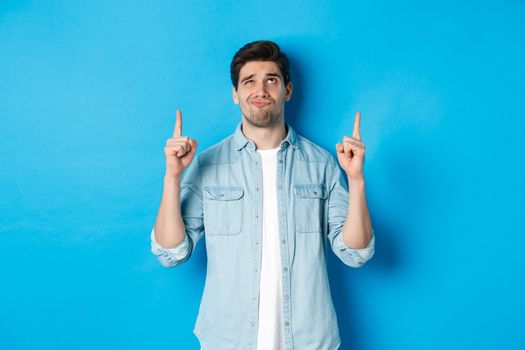 Portrait of doubtful adult man looking disappointed, pointing fingers up at something with uncertain face, standing over blue background.