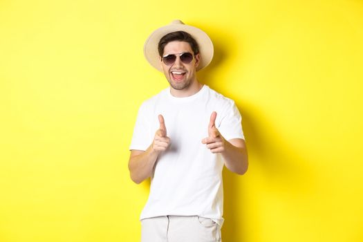 Confident and cheeky guy on vacation flirting with you, pointing finger at camera and winking, wearing summer hat with sunglasses, yellow background.