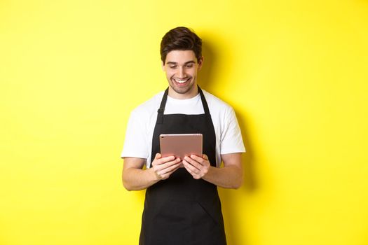 Seller in black apron looking at digital tablet screen, smiling pleased, standing over yellow background.