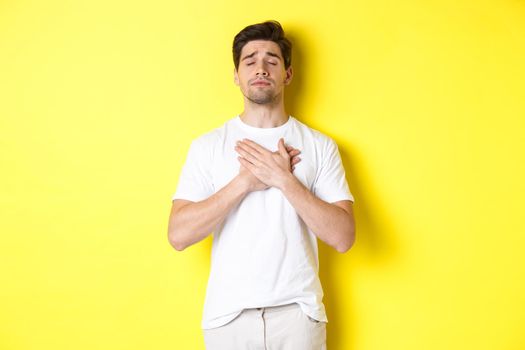 Romantic and nostalgic man holding hands on heart, close eyes and remember something, standing over yellow background.