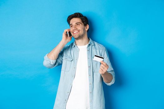 Smiling man call bank support, talking on mobile phone and holding credit card, standing against blue background.