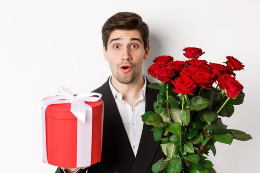 Close-up of attractive man in suit looking surprised, holding gift box and bouquet of roses, giving present for holiday, standing against white background.