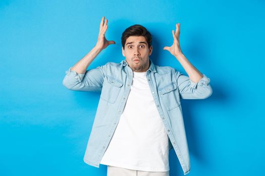 Distressed guy showing mind blowing gesture, looking frustrated and anxious, standing against blue background.