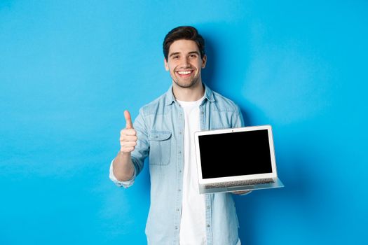 Smiling handsome man showing laptop screen and thumb-up, like promo offer, recommending website, standing against blue background.