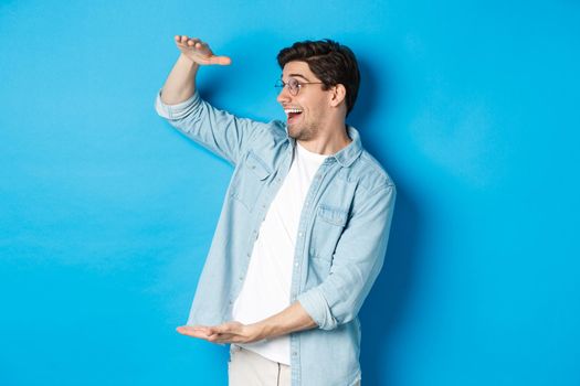 Excited handsome man showing big size object and looking amazed, standing over blue background.