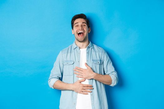 Happy handsome 30 year old man touching belly, laughing out loud and looking at something funny, standing against blue background.