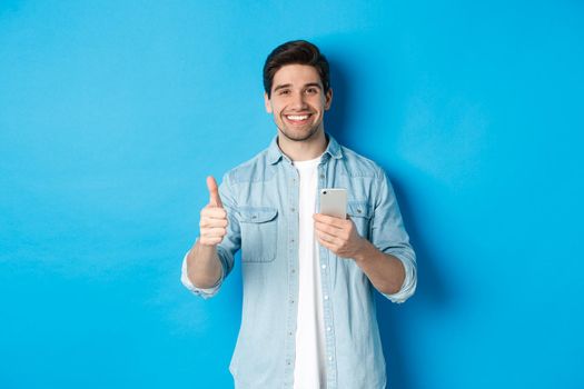 Concept of online shopping, applications and technology. Satisfied man in casual clothes smiling, showing thumbs up after using smartphone app, standing over blue background.