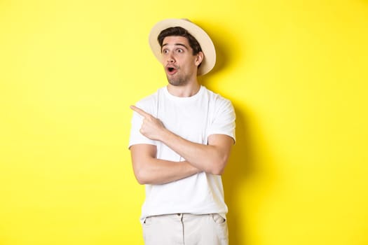 Concept of tourism and lifestyle. Excited handsome guy in straw hat checking out advertisement, pointing and looking at upper left corner logo, yellow background.