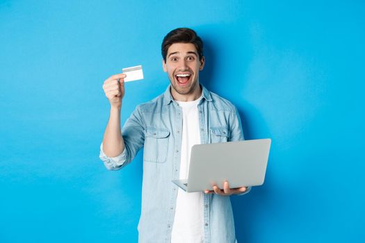 Excited man shop online, showing credit card and holding laptop, buying in internet, standing over blue background.