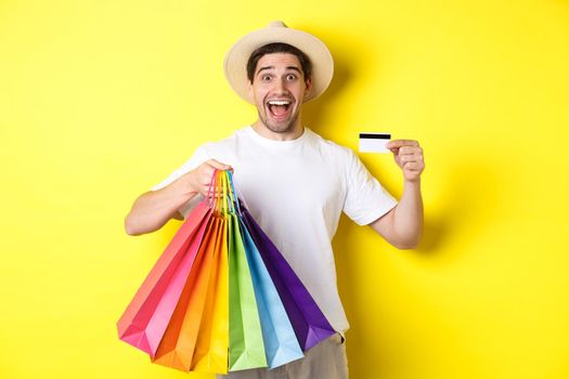 Happy guy going shopping on vacation, holding paper bags and showing credit card, standing over yellow background.