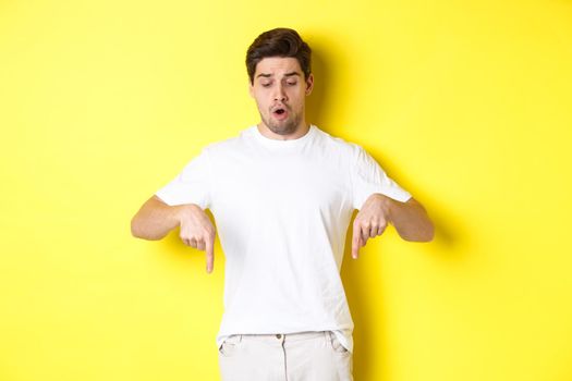 Worried guy pointing fingers and looking down, standing startled against yellow background.