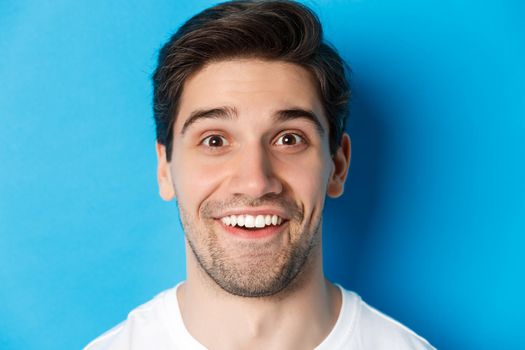 Head shot of handsome male model looking amazed, smiling wondered, standing over blue background. Copy space