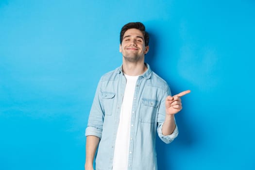 Confident handsome guy smiling sassy, pointing finger right at copy space, showing advertisement, standing over blue background.