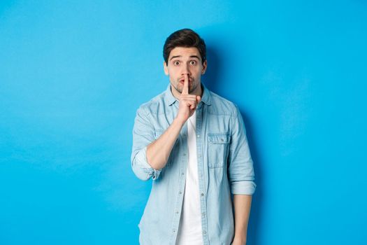 Portrait of excited man asking to keep quiet, showing hush taboo sign and looking nervously at camera, standing against blue background.