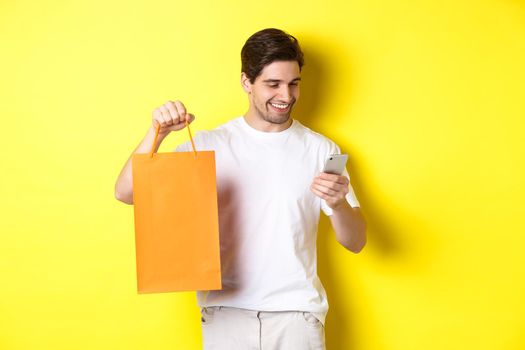 Concept of discounts, online banking and cashback. Happy guy showing shopping bag and looking satisfied at mobile screen, yellow background.